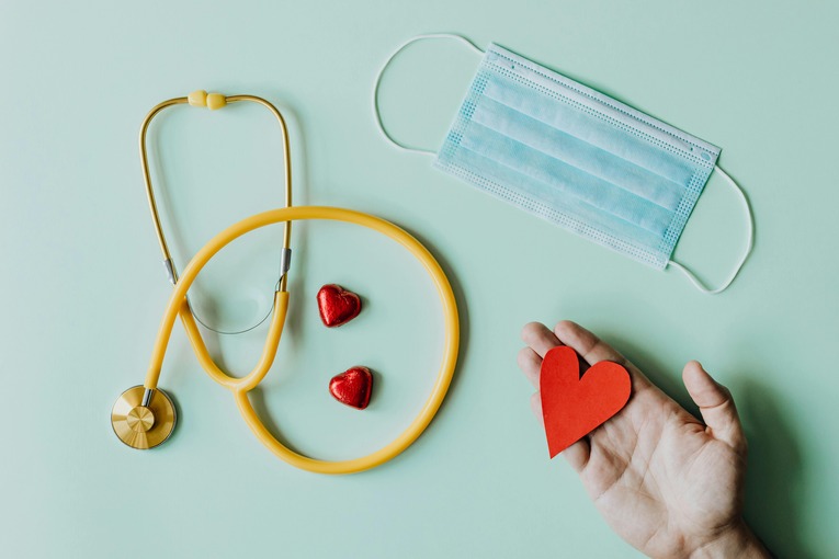 Yellow stethoscope, chocolate hearts, hand holding a red heart, and a blue mask on a light green background.