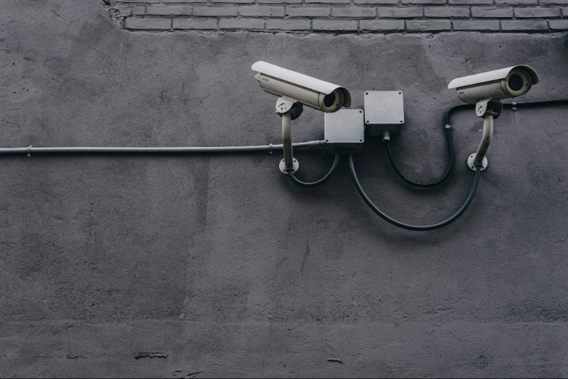 Two surveillance cameras mounted on a gray concrete wall with visible wiring, under a section of brickwork.