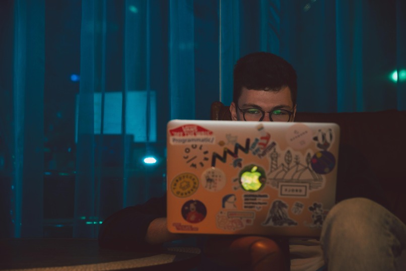 Person with glasses using a laptop covered in colorful stickers, seated in a dimly lit room with a blue curtain in the background.