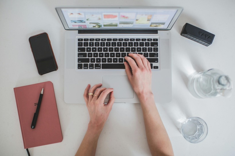 Person typing on a laptop, surrounded by a notebook, smartphone, speaker, and a water bottle on a white desk.
