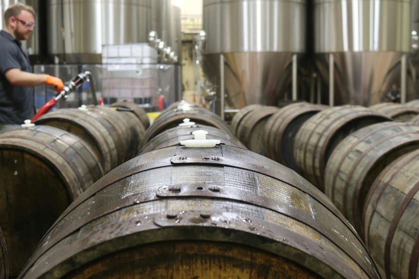 A worker fills wooden barrels with liquid in a brewery, with large stainless steel tanks in the background.
