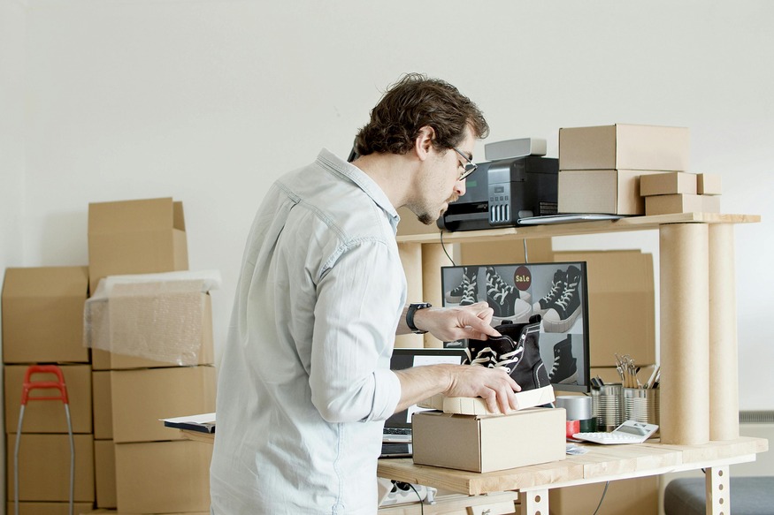 Man assembling a cardboard box at a workbench with stacked boxes, a printer, and a monitor in an office space.