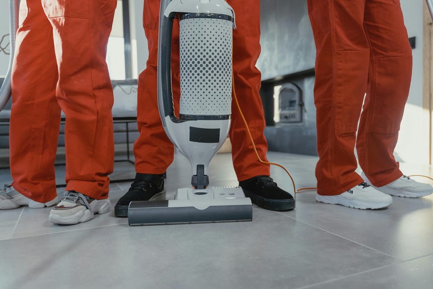 Three people in orange uniforms standing next to an upright vacuum cleaner on a tiled floor.