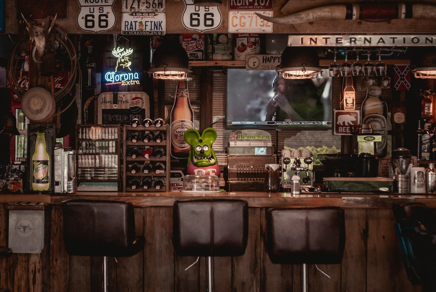 Rustic bar with Route 66 signs, neon lights, a TV, wine rack, and stools. Decor includes vintage items and Corona beer sign.