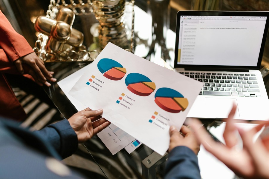 Person holding paper with pie charts near a laptop on a glass table, surrounded by decorative items.