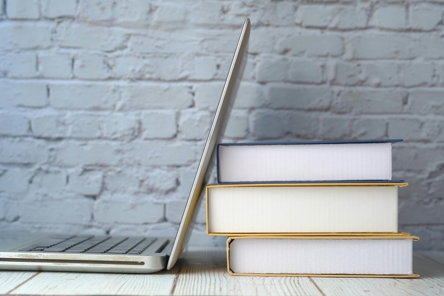 A laptop is partially closed and leaning on a stack of four books against a white brick wall on a wooden surface.