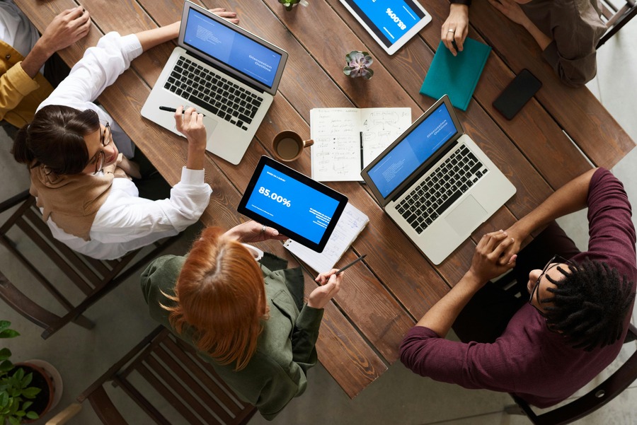 People sitting around a wooden table with laptops and tablets, discussing and taking notes, viewed from above.