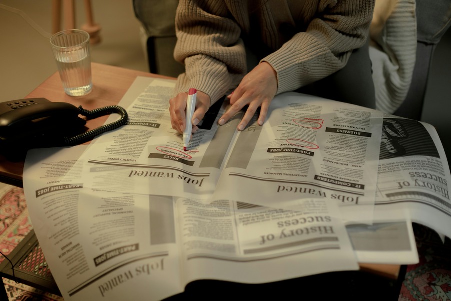 Person in a sweater marking job listings in a newspaper with a red pen; vintage phone and glass of water on the table.