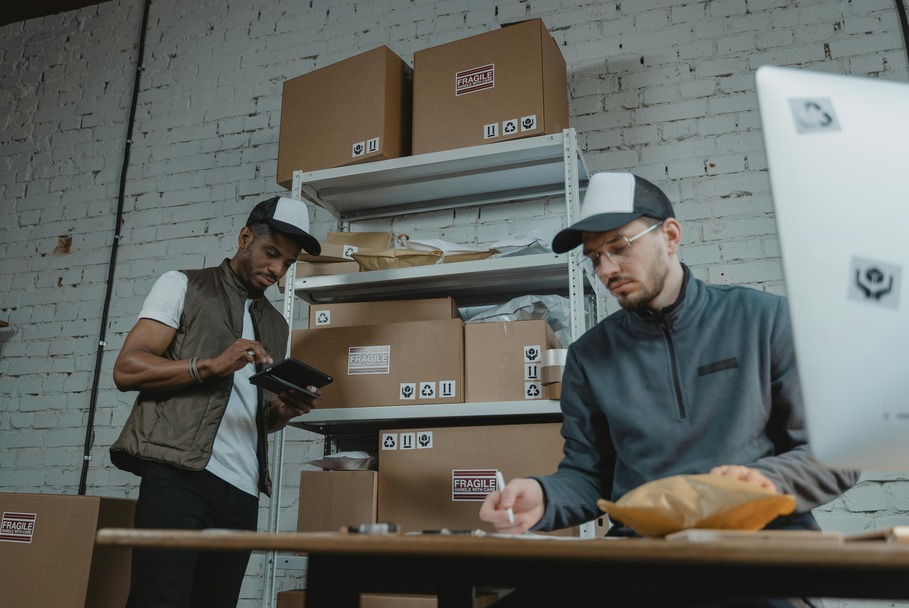 Two workers in caps preparing packages at a packing station with shelves of cardboard boxes.