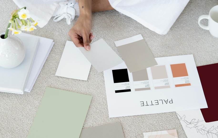 Person arranging color swatches and papers on a carpeted floor with a vase of flowers and a mug nearby.