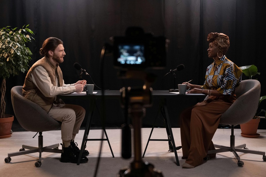 Two people sit at a table in a studio, engaging in a discussion. A camera is set up in the foreground.