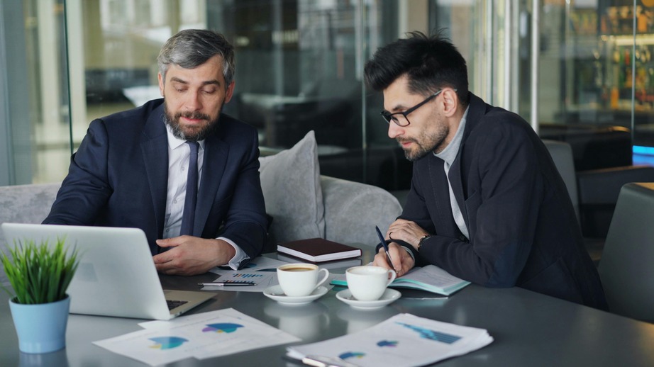 Two men in suits discussing work at a table with a laptop, documents, and coffee cups in a modern office setting.