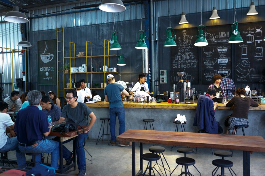 People socialize and work in a modern, industrial-style café with a chalkboard menu and baristas serving drinks behind the counter.