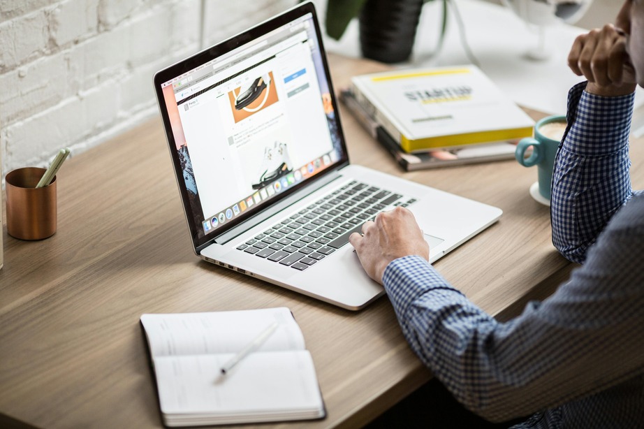 Person using a laptop at a wooden desk with an open notebook, plant, and a blue mug. The screen displays a webpage with a shoe image.