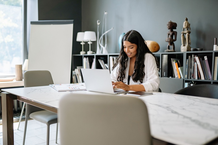 A woman working on a laptop at a marble table in an office with bookshelves and sculptures in the background.