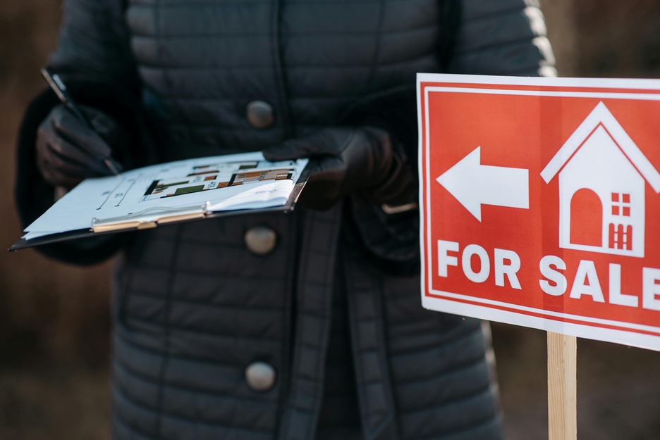 Person in a dark coat holding a clipboard near a red "For Sale" sign with an arrow pointing left.