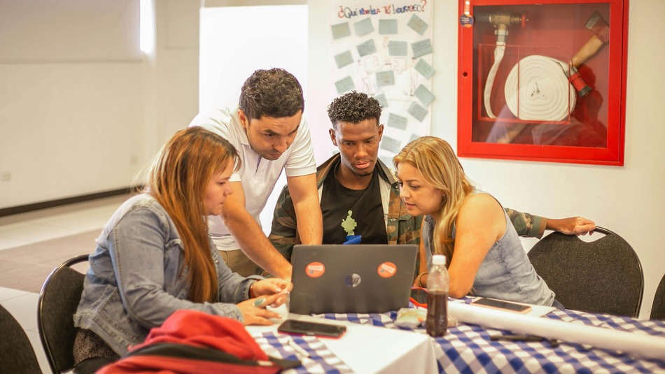 Four people collaborate around a laptop at a table with a checkered tablecloth, focused on the screen in a well-lit room.