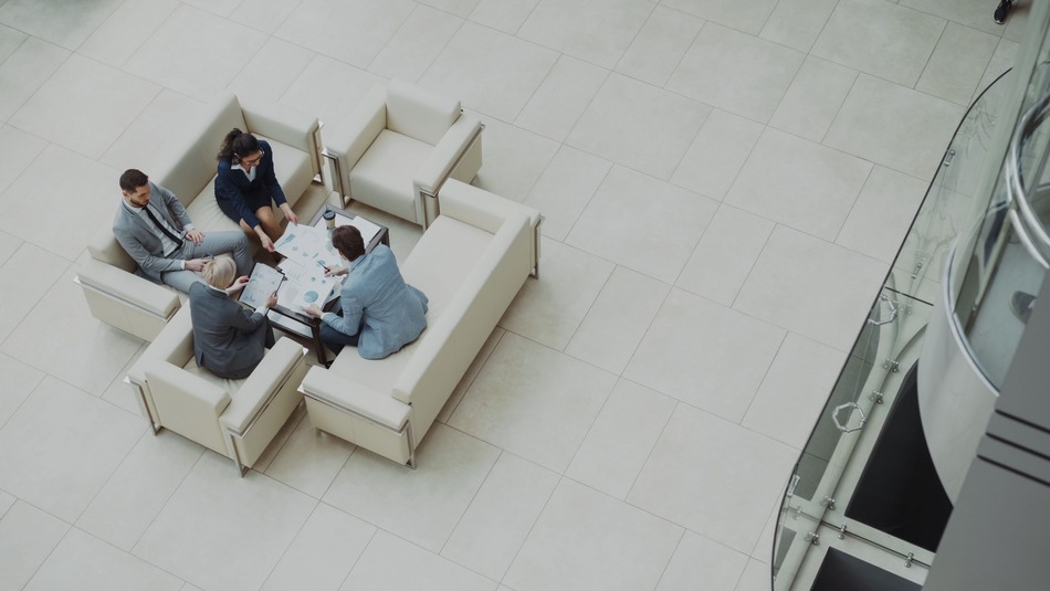 Four people in formal attire seated around a coffee table, discussing papers in a bright, modern office space. Top-down view.