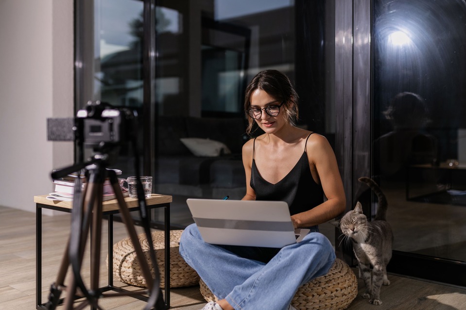 A woman sits cross-legged on a cushion, using a laptop with a camera on a tripod and a cat beside her, in a modern living room setting.