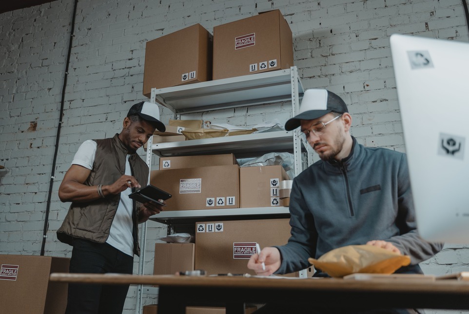 Two workers in caps organize packages in a warehouse with white brick walls and shelves filled with boxes. One uses a tablet, the other labels parcels.