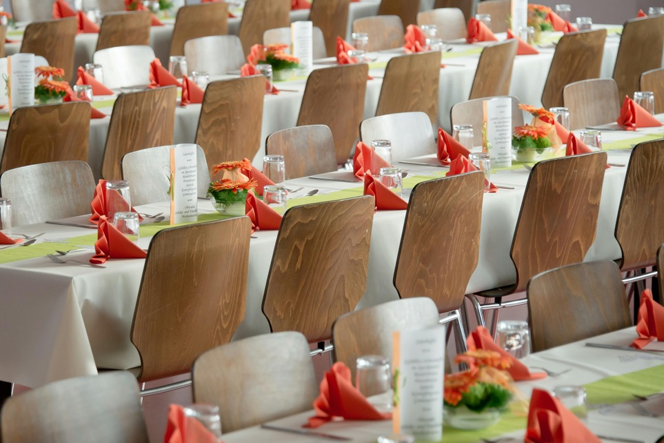 Rows of tables with white tablecloths, wooden chairs, orange napkins, menus, and floral centerpieces, set for an event.