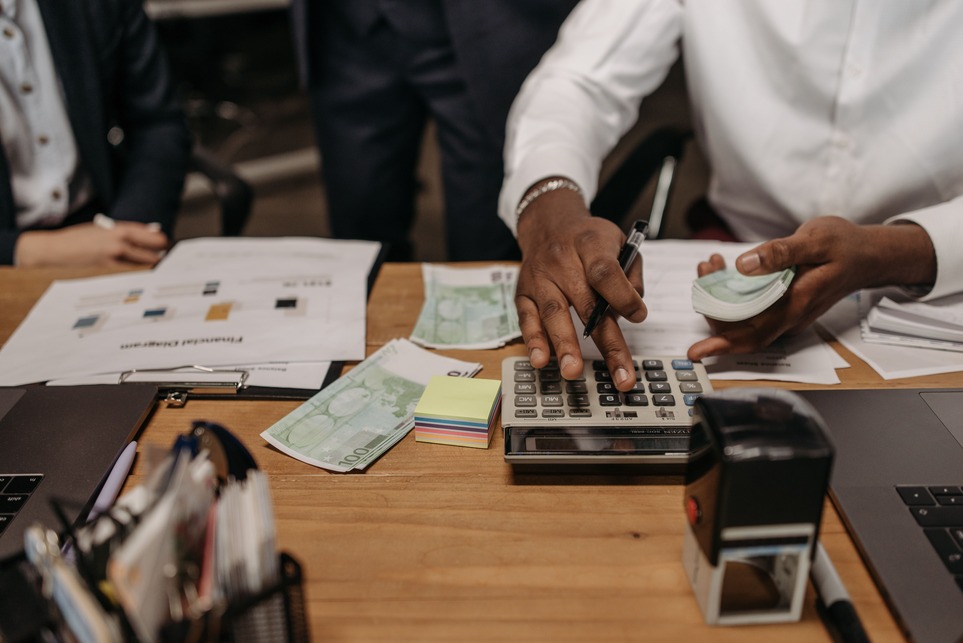 A person uses a calculator on a desk cluttered with cash, papers, and office supplies, while holding a stack of banknotes in the other hand.