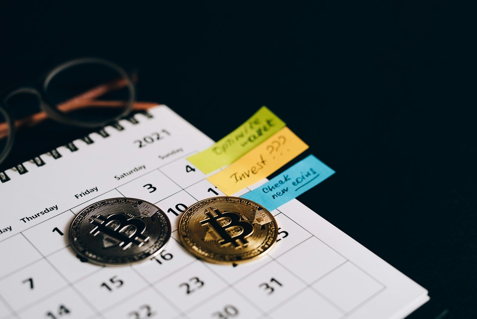 Calendar with Bitcoin coins on a date, surrounded by post-it notes labeled "Invest?", "Check new coins," and glasses nearby.