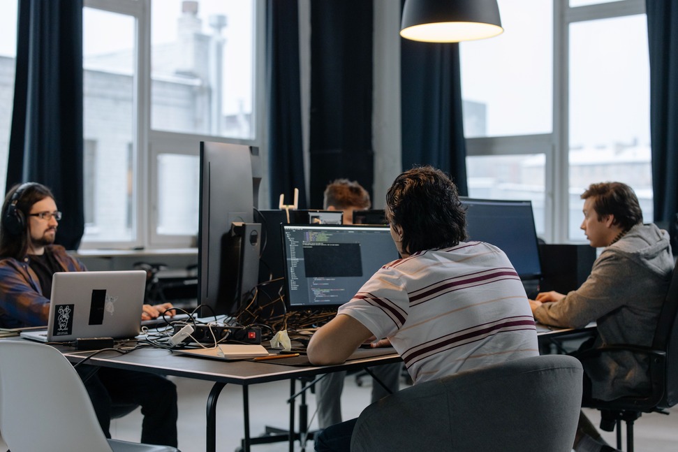 People working on computers in a modern office with large windows and overhead lighting. They appear focused and engaged in their tasks.
