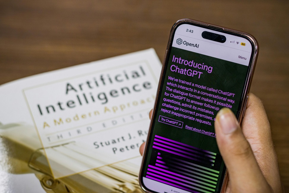 Hand holding a smartphone displaying ChatGPT introduction by OpenAI, with the book "Artificial Intelligence: A Modern Approach" in the background.