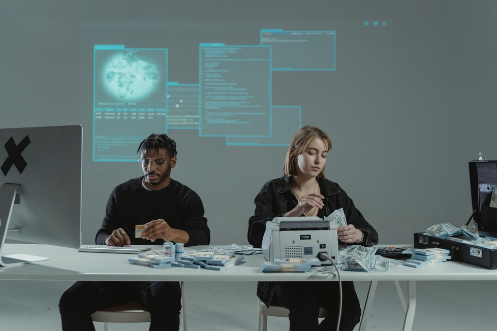 Two people at a table with cash, a money counter, and multiple screens displaying digital data projections in the background.