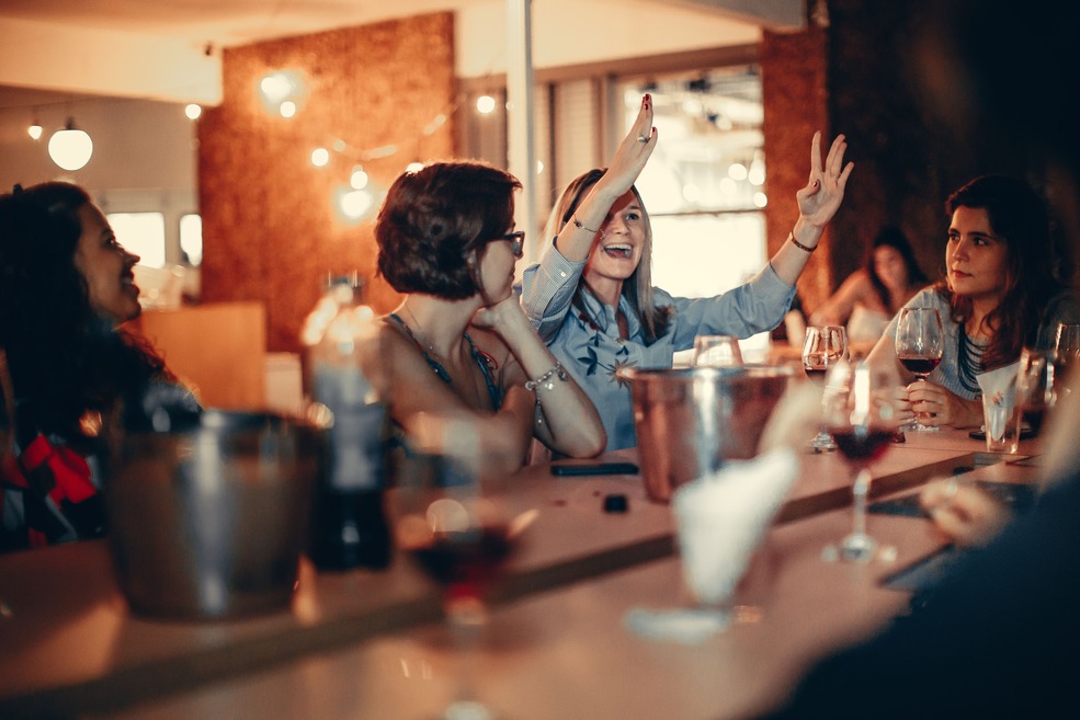 A group of women enjoying drinks and lively conversation at a stylish bar, with warm lighting and wine glasses on the table.