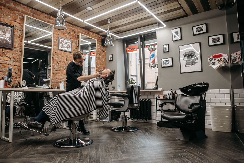 A barber in a modern, stylish barbershop shaves a client's beard. The room features brick walls, framed pictures, and sleek salon chairs.