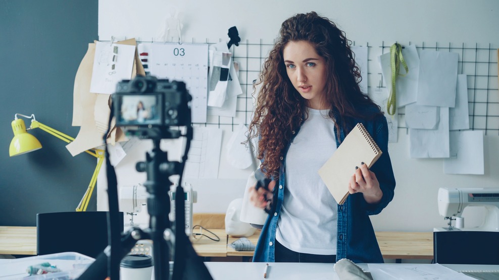 Woman recording a video in a sewing studio, holding a notebook and scissors, with a camera on a tripod and sewing materials in the background.