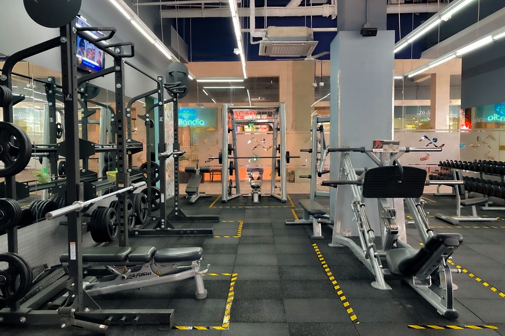 Fitness gym interior with various exercise machines, free weights, and mirrored walls. The space is well-lit with black and yellow flooring.
