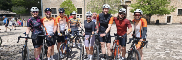 Gruppe von Radfahrern in sportlicher Kleidung posiert draußen mit Fahrrädern vor einem historischen Gebäude bei sonnigem Wetter.