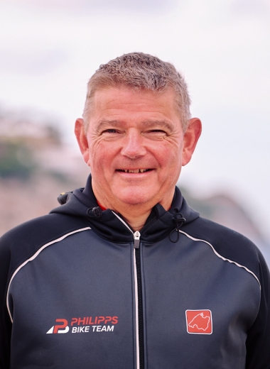 Ein Mann in einer schwarzen Sportjacke mit dem Logo "Philipps Bike Team" lächelt in die Kamera, Hintergrund unscharf.