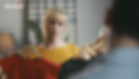 Das Standbild des anklickbaren Videos zeigt eine attraktive Frau in einem knallgelben Kleid, die sich zwei weitere Kleider an den Körper hält und mit fragendem Blick einen Mann anschaut. Sie schaut in unsere Richtung, während wir den Mann nur verschwommen im Anschnitt, von hinten erahnen.