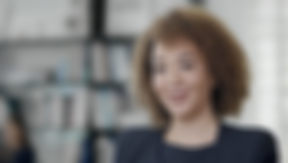 Standbild eines anklickbaren Videos. Interviewsituation. Geschäftsfrau mit Locken grinst in die Kamera. Im Hintergrund ist unscharf eine Bücherwand zu erkennen. Das Bild ist lichtgeflutet.