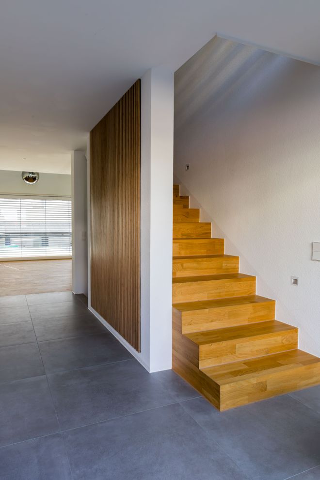 Wooden stairs beside a vertical wood-paneled wall, leading upward in a modern, minimalist room with large windows and gray tile flooring.