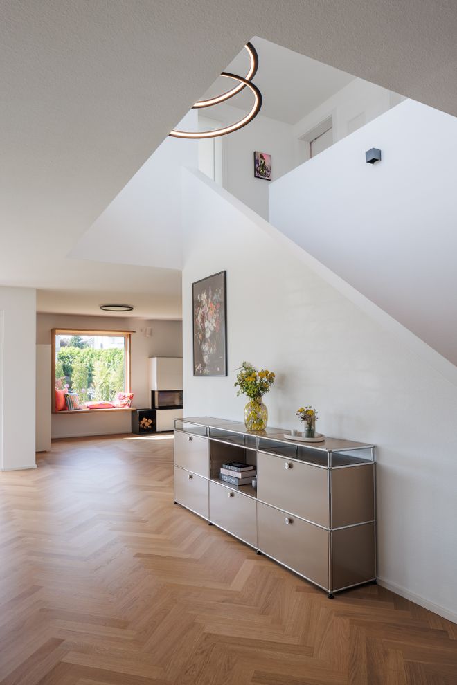 Modern interior with a herringbone wooden floor, sleek metal console, floral artwork, circular ceiling lights, and a cozy window seat with cushions.