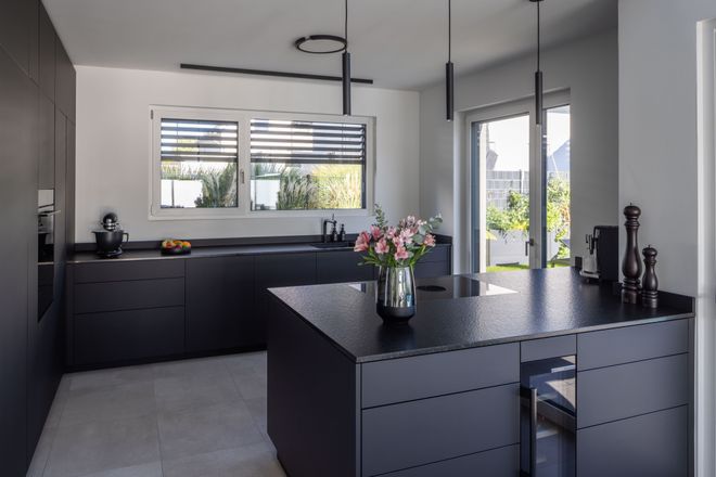 Modern kitchen with black cabinets, large island, and potted flowers. Sunlit window and glass doors with garden view in the background.