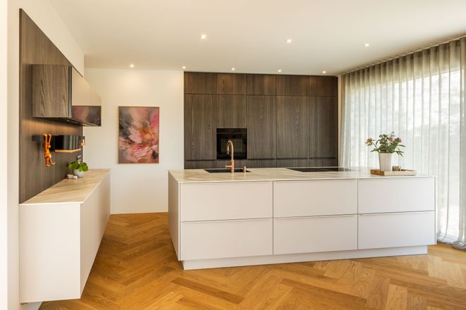 Modern kitchen with a white island, dark wood cabinets, herringbone wood floor, abstract wall art, and large windows with sheer curtains.