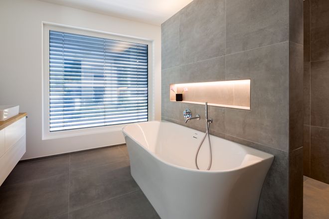 Modern bathroom with a sleek freestanding bathtub, large window with blinds, gray tiled walls, and ambient lighting in a wall niche.