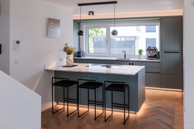Modern kitchen with marble island, three black stools, dark cabinetry, pendant lights, and a large window offering natural light.