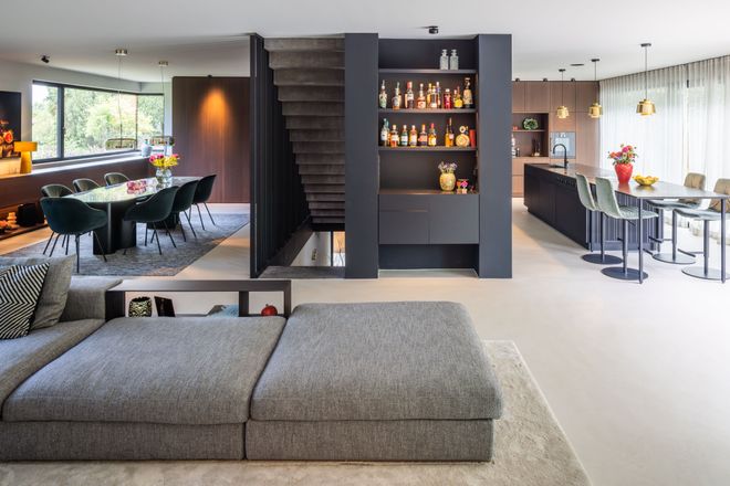 Modern open-plan living area with grey sectional sofa, dining table, bar, and kitchen, featuring sleek decor and large windows.