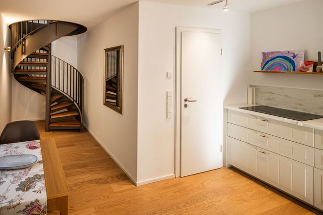 Cozy interior with a spiral staircase, white cabinets, wooden floors, a dining table, and colorful artwork on a countertop.