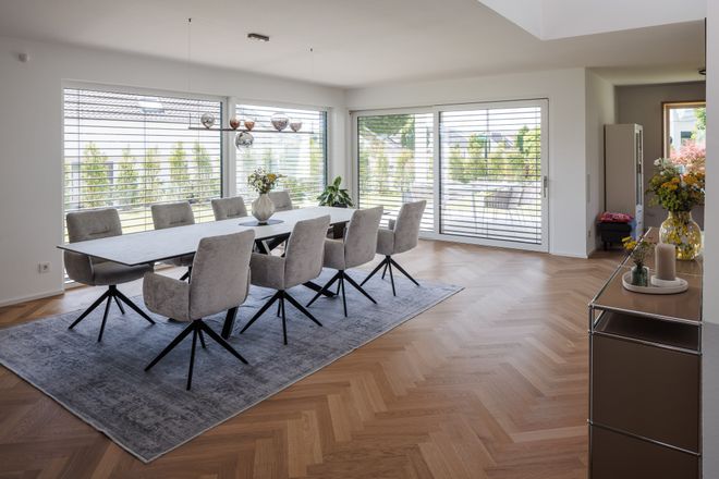 Spacious dining room with a long table, eight beige chairs, herringbone wood floor, large windows, and modern decor.
