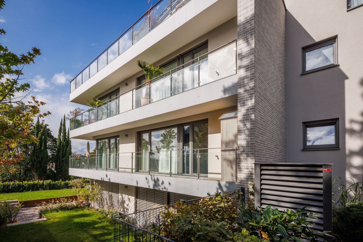 Modern apartment building with three glass balcony levels, surrounded by greenery and basking in sunlight.