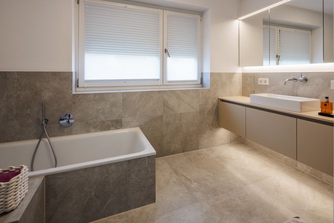 Modern bathroom with a bathtub, large beige tiles, wall-mounted sink, and a lit mirror. Natural light enters through covered windows.