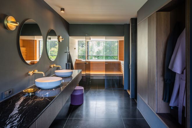 Modern bathroom with dual sinks on a black marble countertop, round mirrors, wall sconces, a wooden bathtub, and a sleek, minimalist design.